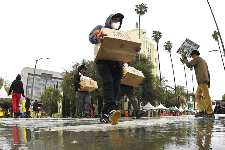 People cross a flooded street with palm trees holding cardboard boxes.