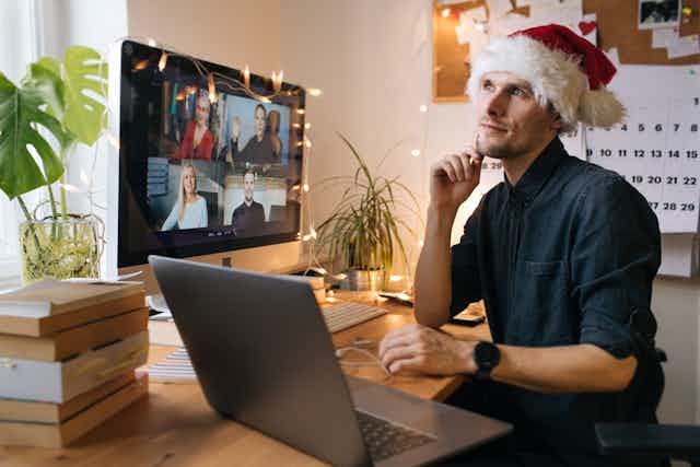 A man sits in a santa hat at his computer with a zoom screen and a laptop in front of him gazing out into the window