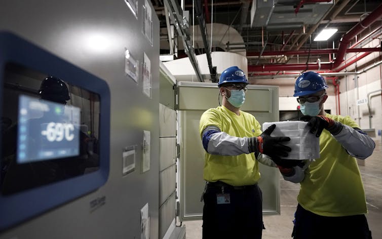 Two workers removing a case of vaccines from an ultracold freezer showing a temperature of minus 69 degrees Celsius