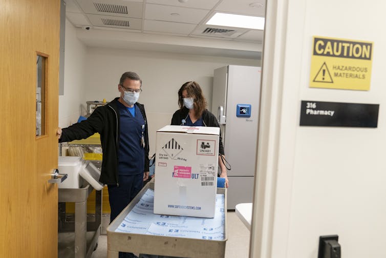 Two pharmacists pushing a cart with a box of Pfizer vaccines.