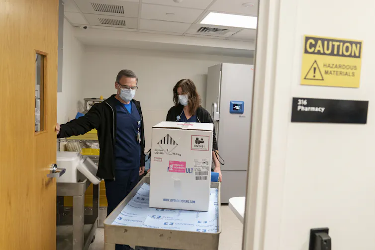 Two pharmacists pushing a cart with a box of Pfizer vaccines.