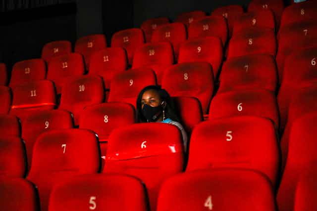 A cinema with red chairs, which are numbered. They are all empty except for one, where a young woman sits wearing a black face mask.