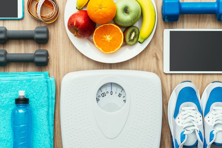 A scale, bowl of fruit, weights, and running shoes, to illustrate weight loss techniques.
