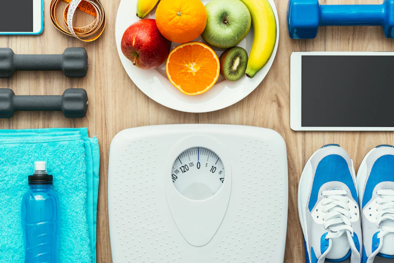 A scale, bowl of fruit, weights, and running shoes, to illustrate weight loss techniques.