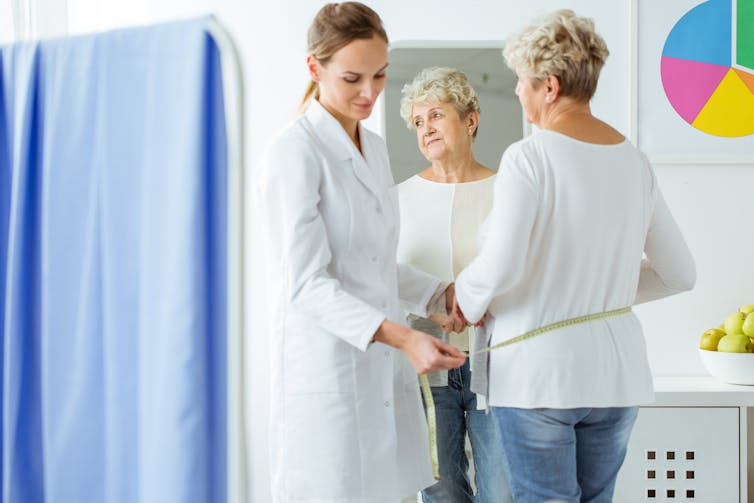 An elderly woman has her waist measured by a female doctor.