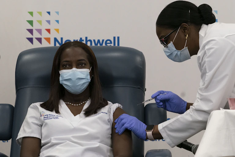 A woman in a mask receives a vaccine from a woman in a labcoat and mask.