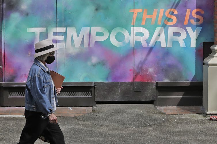 A person wearing a mask walks past a mural on a boarded-up window that reads 'This is temporary.'