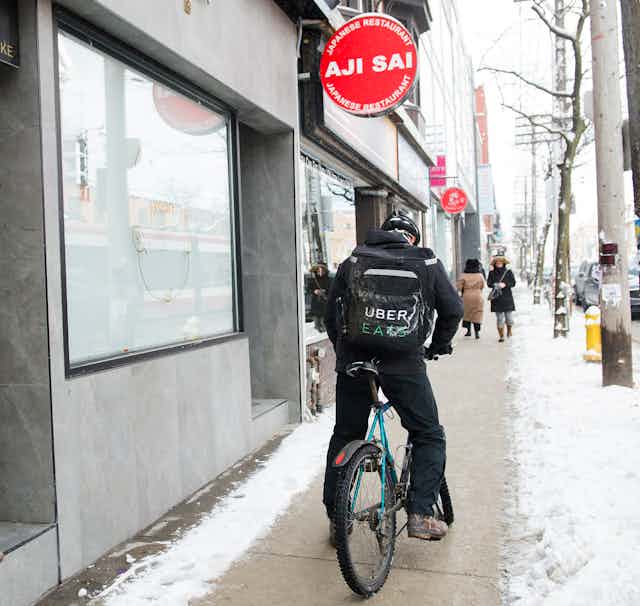 An Uber Eats courier on a bike pick ups an order for delivery on a snowy city street.