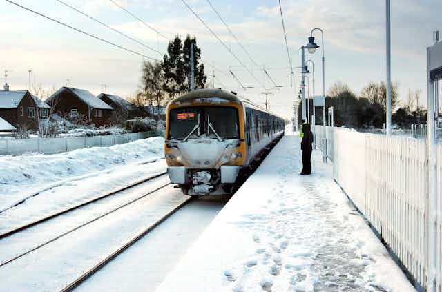 A train pulls into a snowy station with one passenger on the platform.