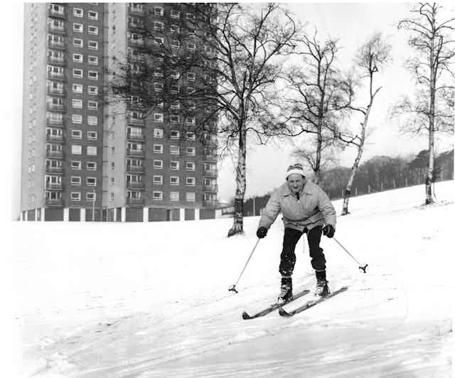 A man in winter attire skis down a snowy hill with a tower block behind him.