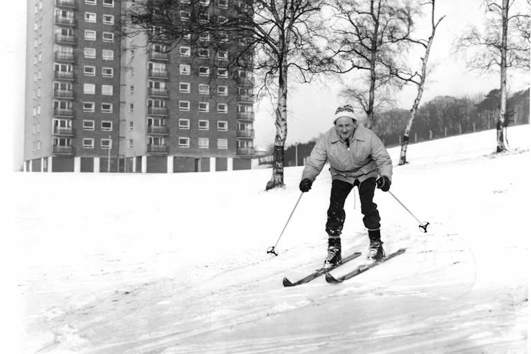 A man in winter attire skis down a snowy hill with a tower block behind him.