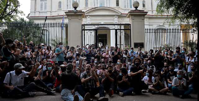 People sit on the ground, clappign and yelling, in front of a large public building