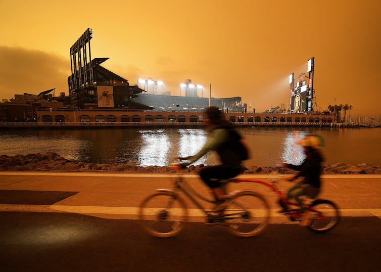 People ride past a ballpark under an orange sky.