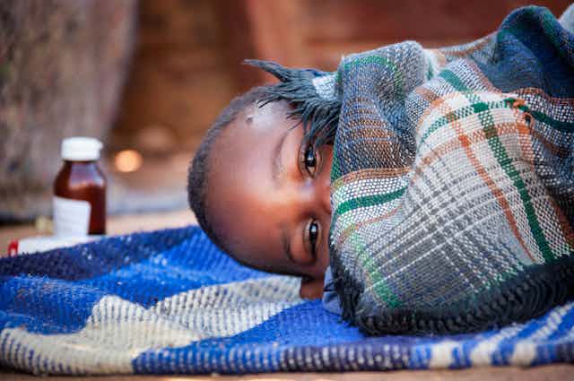 Un niño de Botsuana tumbado, tapado con una manta, mira a cámara. A su lado un bote de medicinas.