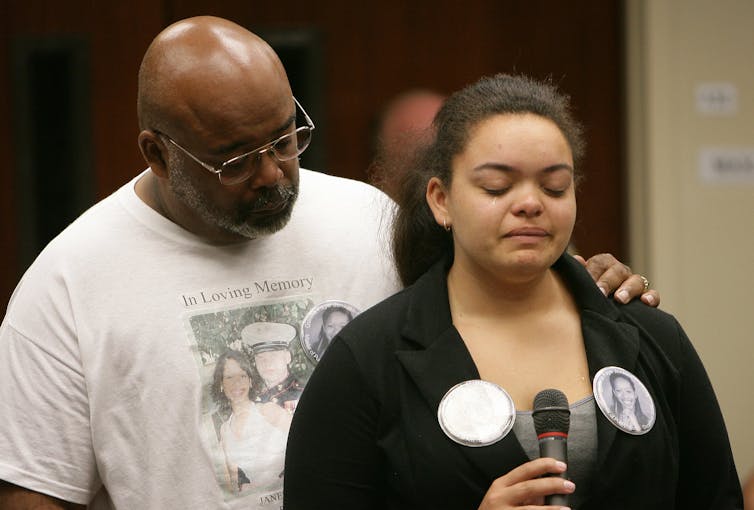 Woman cries in front of a microphone and is consoled by an older man.