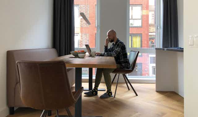Un hombre habla por teléfono ante un ordenador en la mesa del comedor de una vivienda.