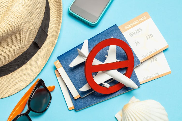 Hat, sunglasses, mobile phone, shell, two passports with boarding passes and a plane figurine with a no symbol over it