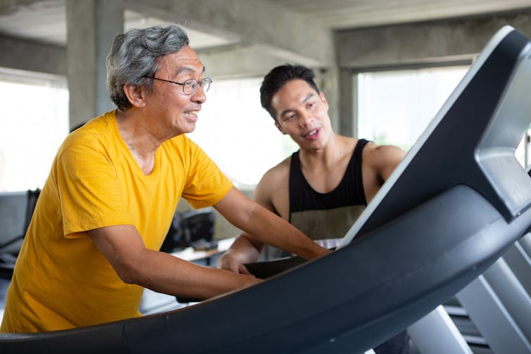 Senior man walking on a treadmill with his young male trainer giving instructions.