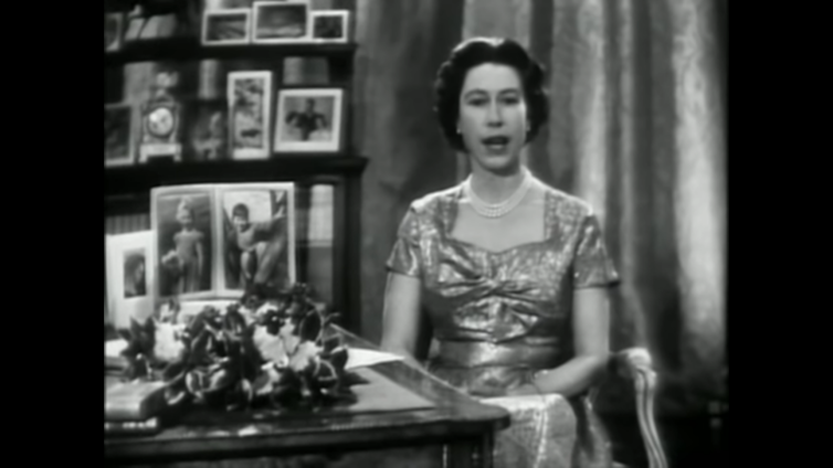 Black and white screenshot of Queen Elizabeth II with framed photos in the background