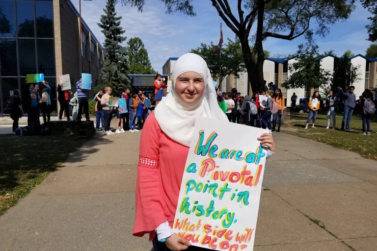 A young woman wearing a hijab holds a sign that says 'We are at a pivotal point in history. What side will you be on?'
