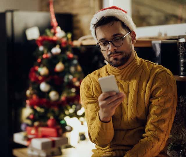 A man sits beside a Christmas tree wearing a santa hat with his phone in his hand, looking sad