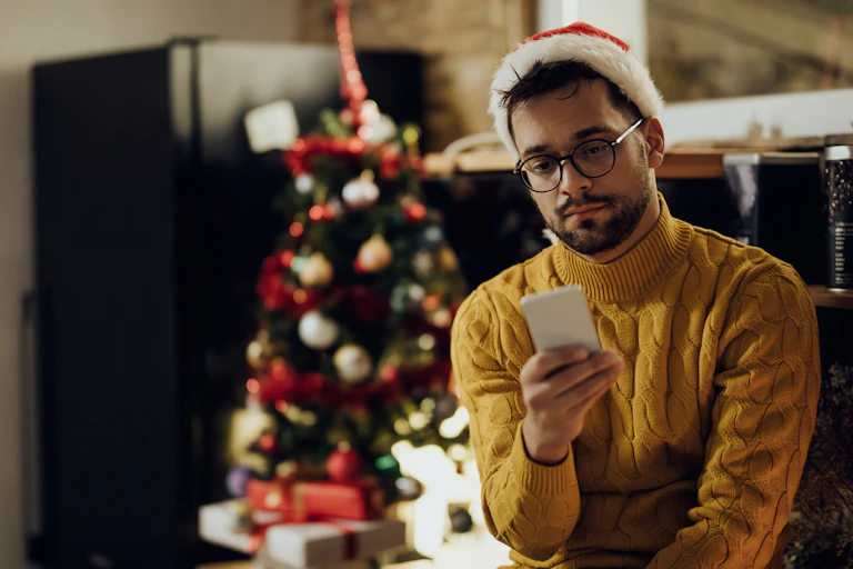 A man sits beside a Christmas tree wearing a santa hat with his phone in his hand, looking sad