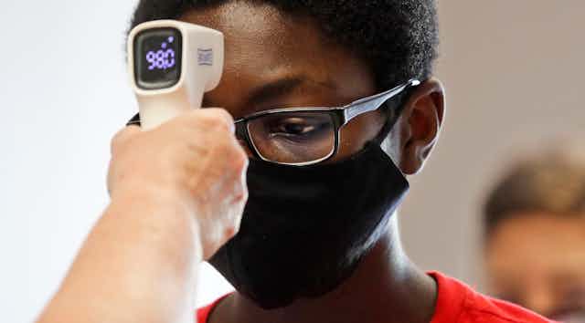 A hand holds a device with a display reading 98.0 to the forehead of a boy who is wearing glasses and a facemask