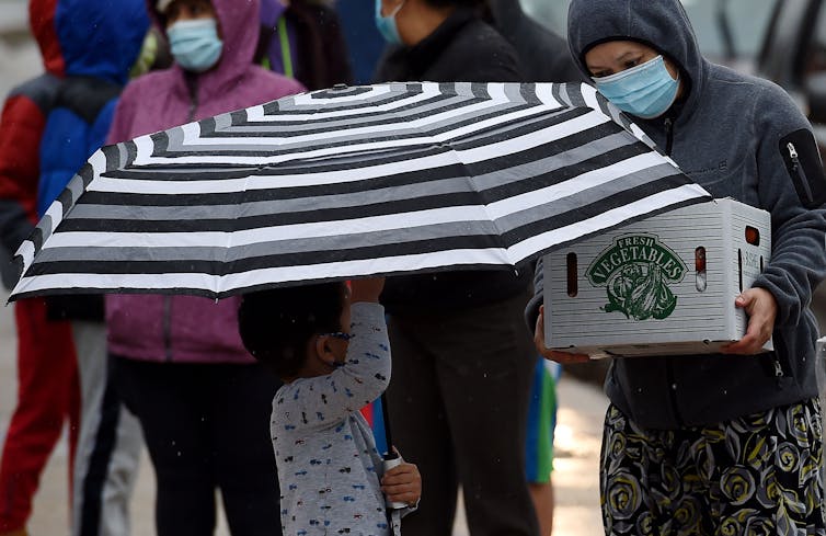 A small child holds a big umbrella while their mom in a mask holds a box that says 'vegetables.'