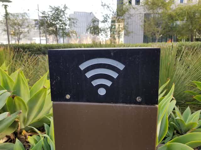 A black box with the Wi-Fi symbol on one side on a brown pedestal amidst plants in a city park