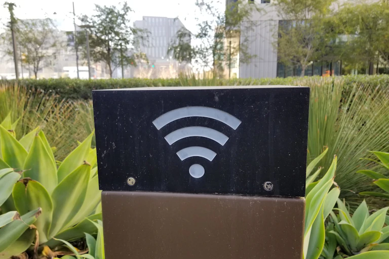 A black box with the Wi-Fi symbol on one side on a brown pedestal amidst plants in a city park