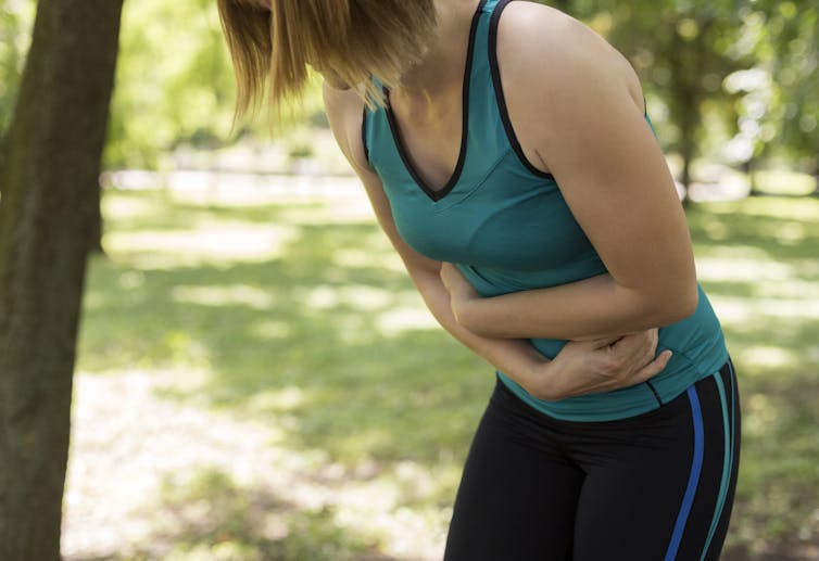 A woman in exercise clothes is doubled over, holding her stomach