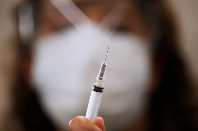 A blurred woman in the background holds up a syringe containing COVID-19 vaccine.