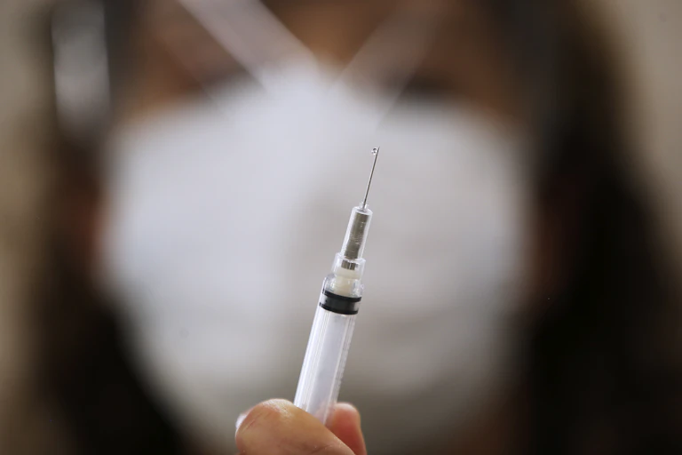 A blurred woman in the background holds up a syringe containing COVID-19 vaccine.