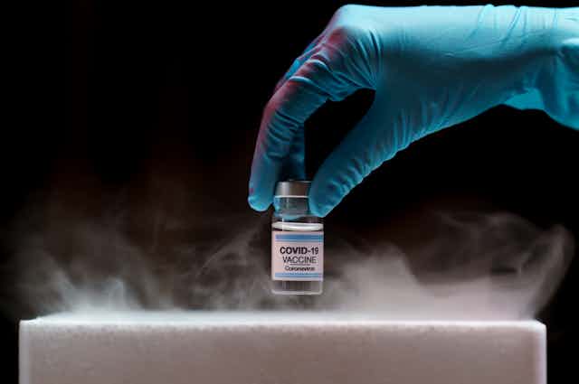 Una mano con guante azul saca un bote de vacuna covid-19 de un receptáculo frigorífico.