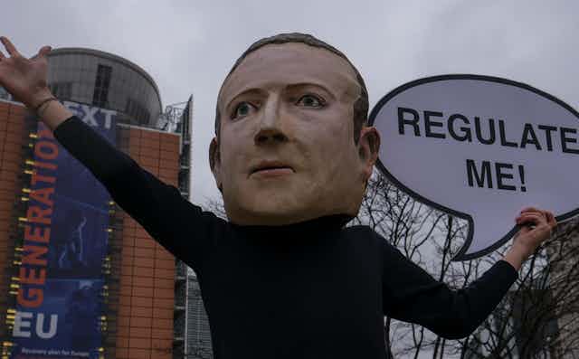 A person in an oversized mask of Mark Zuckerberg holds up a sign outside the European Commission offices in Brussels in December 2020 with a sign saying "regulate me"