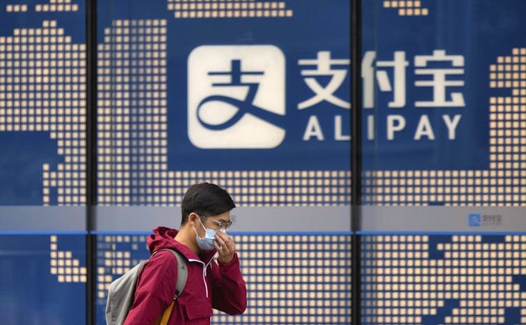 A person in a mask walks past a digital screen of an Alipay sign