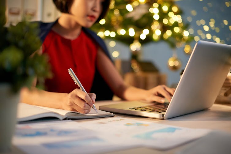 Woman writing in notepad with one hand and typing on laptop with the other in front of Christmas tree