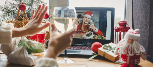 Person raising a glass of wine and waving to a woman wearing a Santa hat and drinking wine on a video call on a laptop