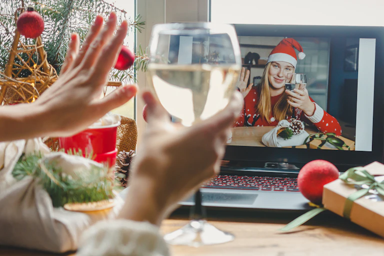 Person raising a glass of wine and waving to a woman wearing a Santa hat and drinking wine on a video call on a laptop