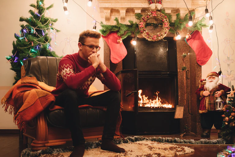 A man sits in a chair next to a fireplace surrounded by Christmas decorations.