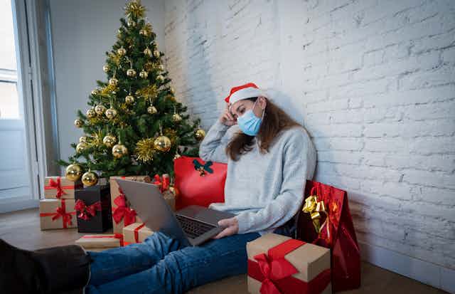 Sad woman alone with computer, mask and christmas tree