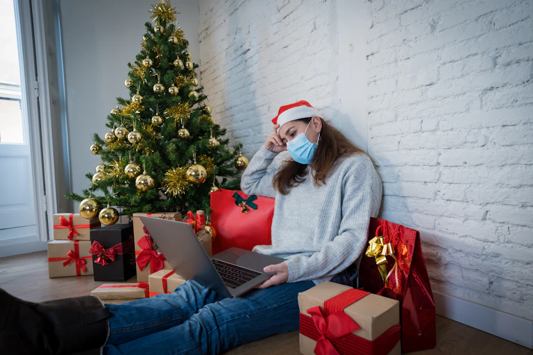 Sad woman alone with computer, mask and christmas tree