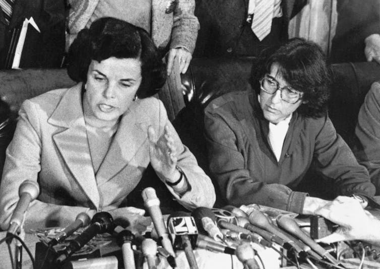 Black and white image of Feinstein speaking into several microphones, seated