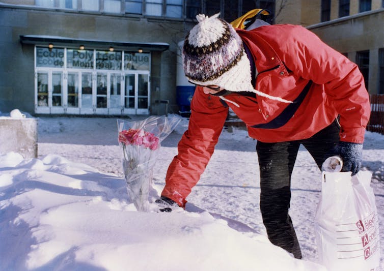 A man in a snow hat and red ski jacket places a bouquet of roses in a snowbank outside a school.