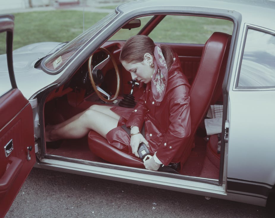 A 1960's era woman is fastening her seatbelt in a sporty car.