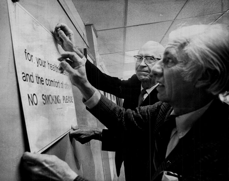 Dos políticos hombres mayores de la década de 1970 colocan un letrero que dice "Por su salud y seguridad y la comodidad de los demás, no fumar".
