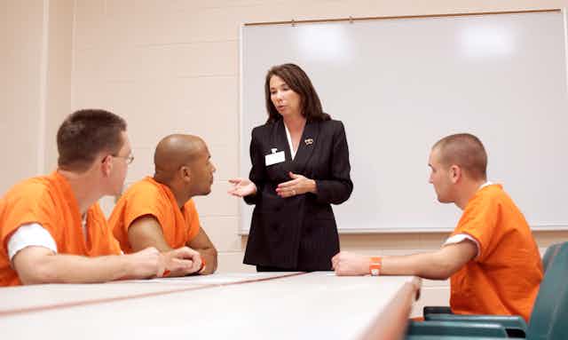 A woman stands at the front of a room with a whiteboard as three prisoners in orange jumpsuits pay attention