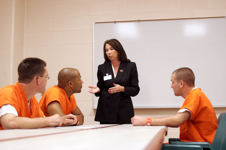 A woman stands at the front of a room with a whiteboard as three prisoners in orange jumpsuits pay attention