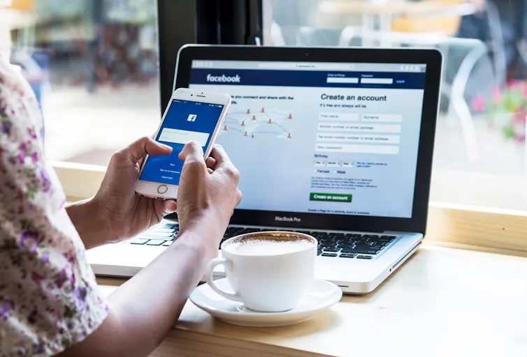 Person looking at Facebook on their phone and on their laptop with a cup of coffee next to them