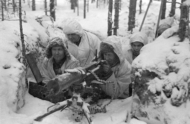 Five Finnish soldiers in winter gear man a machine gun nest surrounded by snow.